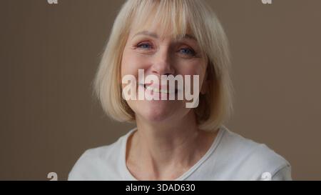 Happy old Caucasian smiling 60s woman smiling toothy carefree joyful pleased looking at camera posing in beige studio indoors cheerful senior female Stock Photo