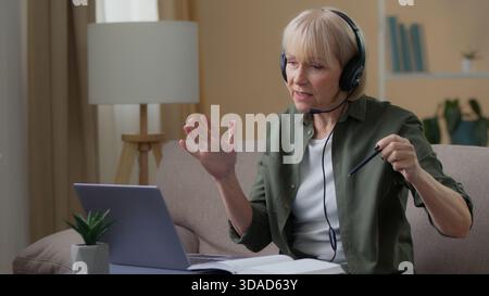 Caucasian old female in headset remote teacher talking online class lesson from home with laptop senior woman talk distant education explaining Stock Photo