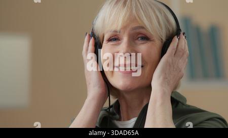 Old Caucasian woman in headphones smiling listening music in headphones looking at camera senior female retired lady happy listen audio grandmother in Stock Photo