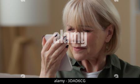 Old Caucasian woman at home trying to see looking in mobile phone screen bad vision blurry eyesight without glasses senior lady aged retired female Stock Photo