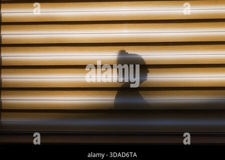Profile silhouette of a woman with tied-up hair cast on a yellow-striped window blind under strong sunlight. Stock Photo