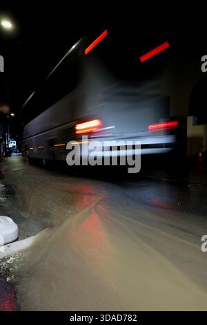 Motion-blurred public bus driving through floodwater at night. A dramatic cityscape with reflections and light streaks. Stock Photo