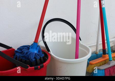 Closeup of household cleaning tools including two buckets with mops and a pair of colorful brooms leaning against a white wall. The vibrant handles an Stock Photo