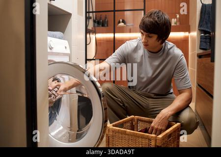 Young Man Loading Laundry into Washing Machine in Modern Bathroom Stock Photo
