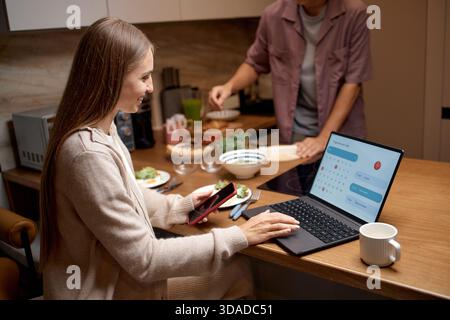 Young Woman Scheduling Appointment on Laptop While Sitting in Kitchen Stock Photo