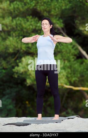 Adult female with dark hair performs focused yoga pose on Sandy Beach near Baltic Sea. Calm expression and natural green forest setting emphasize mind Stock Photo