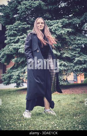 Young woman with long hair stands on grass, wearing dark trench coat and casual shoes, smiling gently with serene pine tree backdrop. Outdoor scene ra Stock Photo