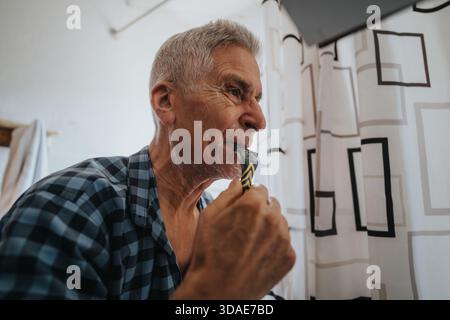 Older man shaving with razor in bathroom wearing plaid shirt, enjoying a morning routine Stock Photo