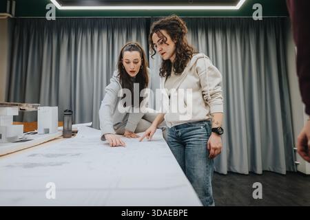 Two women collaborate on architectural plans at a modern studio workspace during a project meeting Stock Photo