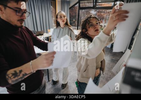 Three students collaborate in a modern classroom, designing a project and presenting ideas on a whiteboard Stock Photo