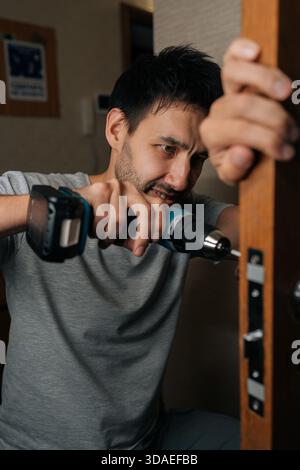 Bearded handyman installing new door lock into wooden door frame with electric screwdriver, focusing on task of home repair and improvement. Concept Stock Photo