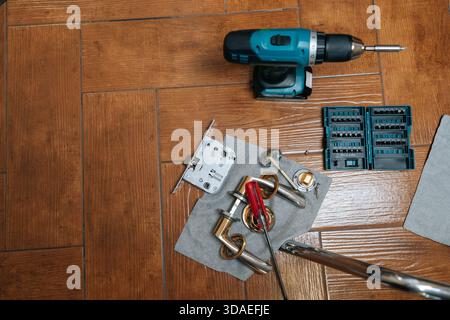 Top view of tools and hardware for door lock installation or repair project, including disassembled handle, latch mechanism, power drill, and Stock Photo