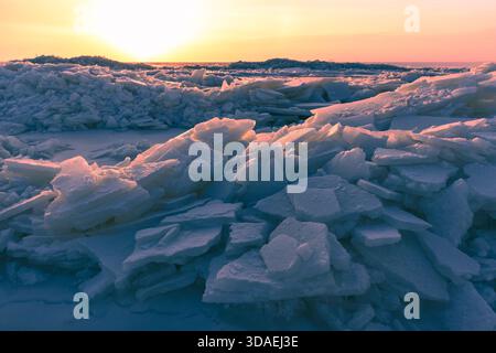 A dramatic winter scene shows broken ice chunks piled along a frozen sea, bathed in warm sunset colors. The blue shadows contrast with orange light, c Stock Photo