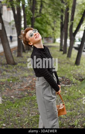 A cheerful middle-aged woman enjoys a walk in a tranquil park, surrounded by lush green trees. She smiles warmly, radiating happiness and contentment Stock Photo