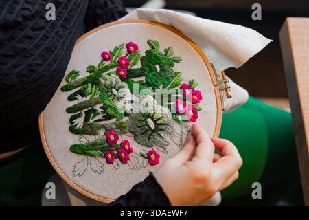 A close-up of hands skillfully embroidering vibrant flowers on fabric using a wooden hoop. The detailed stitches and colorful threads bring the floral Stock Photo