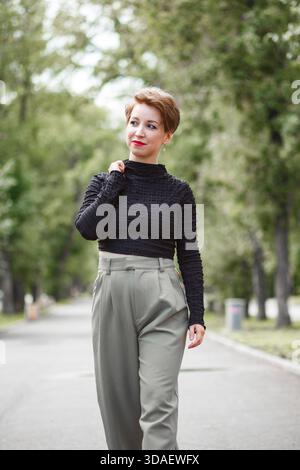 A middle-aged woman with a bright smile is enjoying a leisurely walk in a lush park. Her content expression and confident stride convey happiness and Stock Photo