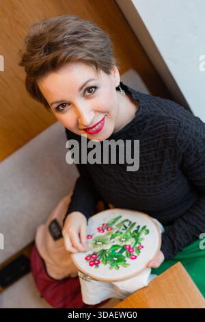 A middle-aged woman, radiating happiness, focuses on her embroidery project. Her cheerful expression captures the joy in crafting and creativity durin Stock Photo