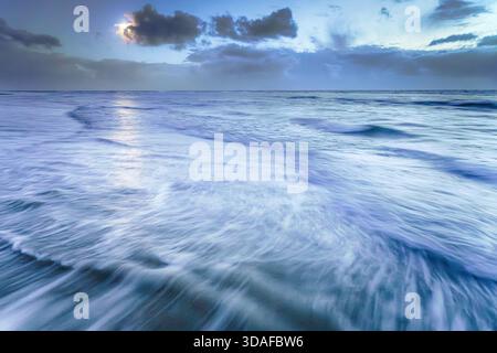 A long exposure shot captures the ethereal movement of waves under a dramatic twilight sky on Ameland. The soft blue hues and blurred water create a s Stock Photo
