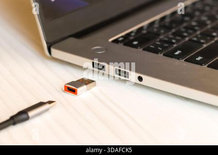 Detailed view of a metallic USB adapter with orange accent next to a USB-C cable, positioned near silver laptop's ports on a white wooden background. Stock Photo