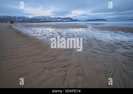 The sandy beach in Teriberka shows patterns in wet sand as waves come in. Mountains covered in snow rise in the background under cloudy skies. Stock Photo