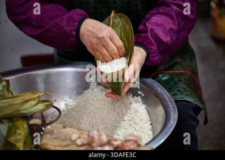 Hands pouring steamed glutinous rice into bamboo leaf cone, traditional dumpling making process detail Stock Photo