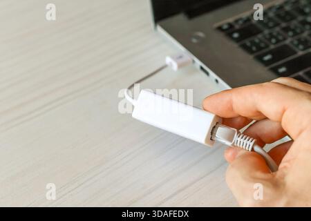 Close-up of a hand connecting a white Ethernet cable to a USB Ethernet adapter plugged into a silver laptop on a light desk, illustrating quick wired Stock Photo