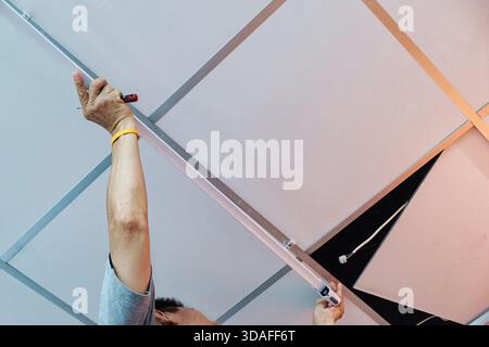 Technician man installing a ceiling mounted light fixture using screwdriver, reaching up with exposed wire and suspended panel Stock Photo