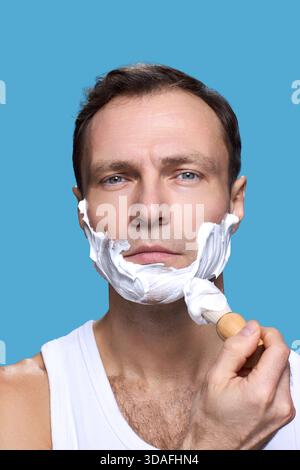 Portrait of Caucasian middle aged man looking into camera applying shaving cream to face with shaving brush against blue background showing daily grooming routine Stock Photo