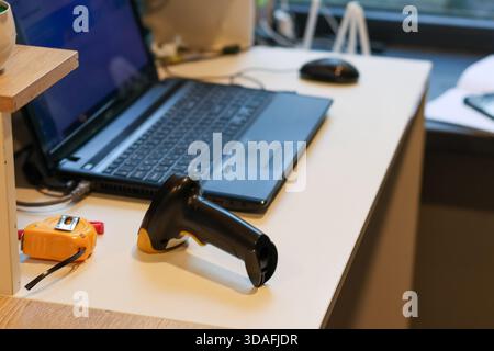 Foreground focus on black and yellow barcode scanner resting on light desk surface with laptop, mouse and tape measure in office setting Stock Photo