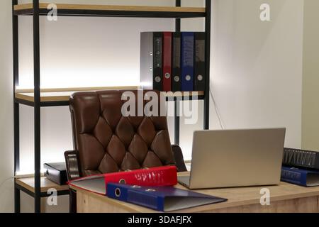 Wooden desk features open silver laptop, colorful ring binders, and brown leather chair against white wall with illuminated shelf holding more folders Stock Photo