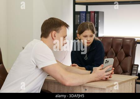 Adult and child share grip on mobile device at office table surrounded by leather chairs and file shelves Stock Photo