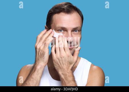 Portrait of middle aged Caucasian man smiling and applying under eye patch to face while looking into camera, demonstrating skincare routine against blue background Stock Photo