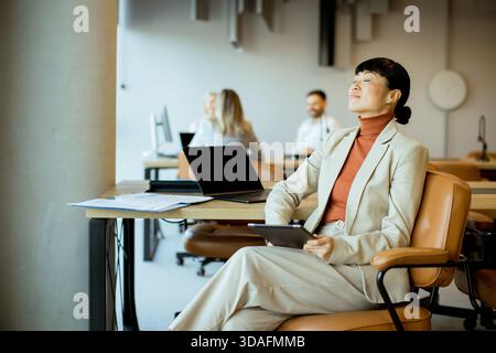 A businesswoman sits relaxed at her desk in a contemporary office, enjoying a moment of tranquility while holding a tablet. Stock Photo