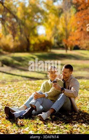In a vibrant park adorned with autumn leaves, a Korean woman and Caucasian man sit closely, sharing smiles and laughter while using a smartphone. Stock Photo
