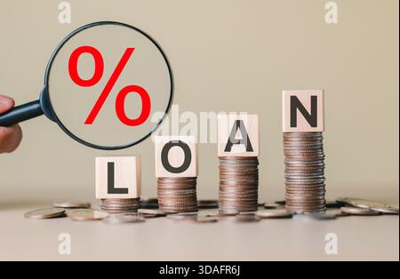 Stacked coins with wooden blocks spelling "LOAN" and a magnifying glass showing a red percentage symbol, representing interest rates, personal finance Stock Photo