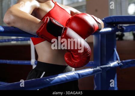 Young female boxer rests on ropes in boxing ring wearing red gloves during training with strong focused attitude Stock Photo
