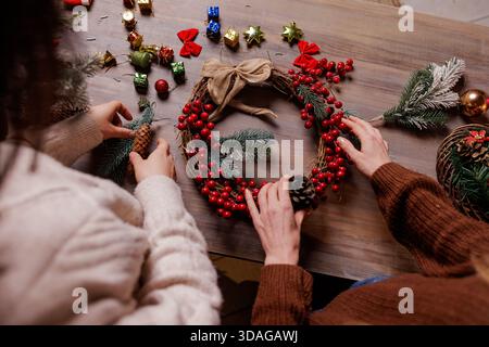 Overhead shot of two women arranging Christmas wreath with red berries, fir sprigs and pinecones on wooden table surrounded by ornaments. Stock Photo