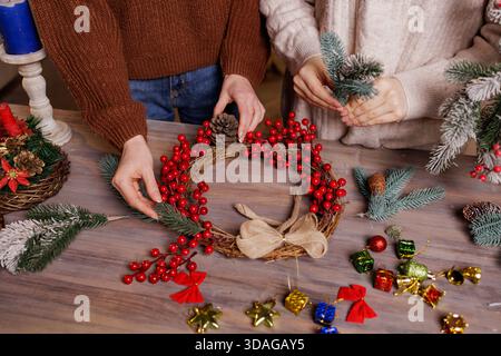 Closeup of two women arranging natural Christmas wreath with red berries, pine branches and cones on rustic wooden table indoors. Stock Photo