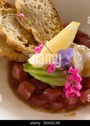 Beautifully decorated tuna tartare dish with avocado and scallop slices, lemon, bread chips and decorated with flowers on a white plate Stock Photo