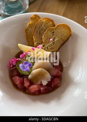 Beautifully decorated tuna tartare dish with avocado and scallop slices, lemon, bread chips and decorated with flowers on a white plate Stock Photo