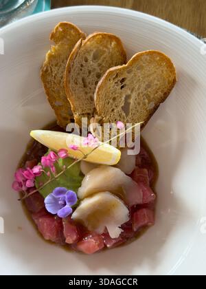 Beautifully decorated tuna tartare dish with avocado and scallop slices, lemon, bread chips and decorated with flowers on a white plate Stock Photo