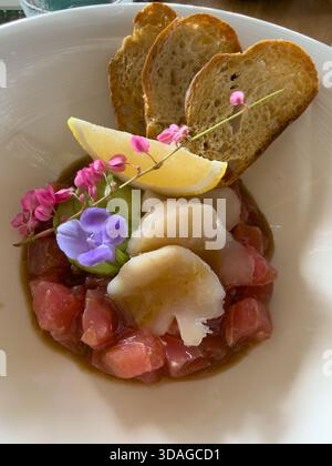 Beautifully decorated tuna tartare dish with avocado and scallop slices, lemon, bread chips and decorated with flowers on a white plate Stock Photo