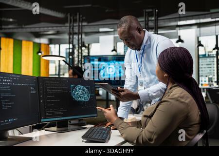 Supervisor overseeing intern using computer in workplace to do maintenance on neural networks. Team leader supervising trainee in tech startup office using AI deep learning systems on PC Stock Photo
