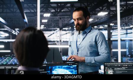 Manager supervising work of server room engineer using AI to review infrastructure diagnostics on devices. Tech company supervisor overseeing IT expert in data center analyzing data using AI, camera A Stock Photo