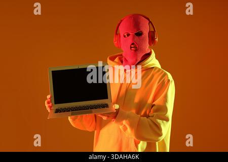 Young man in balaclava with headphones and laptop on yellow background Stock Photo