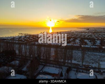 Dramatic aerial view of the bright sun setting over a large lake, creating a vibrant orange glow over the water and illuminating the snow-covered reso Stock Photo