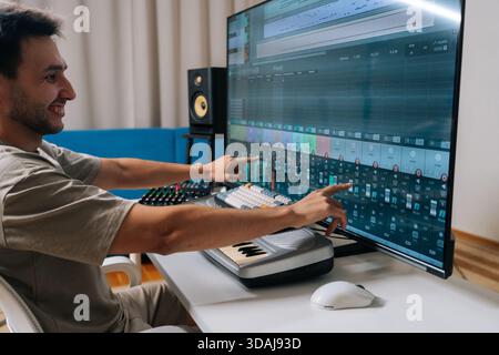Cheerful sound engineer creating new music in home studio, arranging tracks and mixing audio levels with midi keyboard and software on a large display Stock Photo