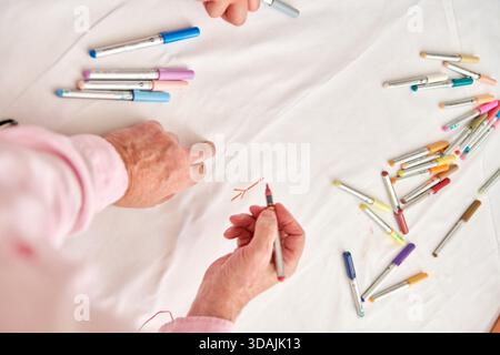 Elderly hands actively sketching on white cloth surface using vibrant markers scattered around. Soft lighting enhances texture and warm tones in creat Stock Photo