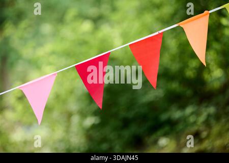 Bright pink, red, and orange triangular pennant flags are strung on white cord against blurred natural environment of vibrant green foliage in dayligh Stock Photo