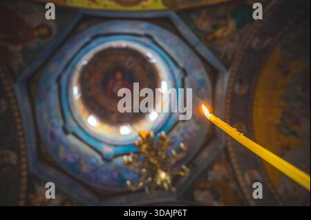 Petrosani, Romania November 22 2025: Lit prayer candle brightly burning flame contrasted against the ornate painted ceiling dome of an Orthodox church Stock Photo